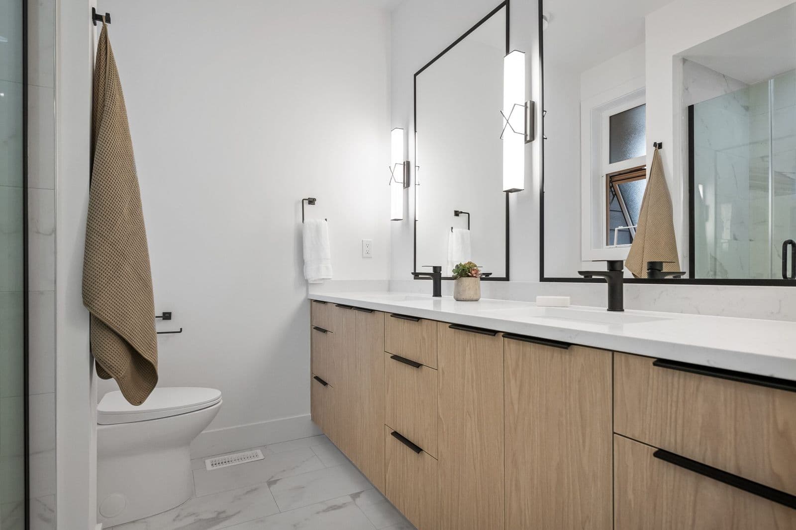 Archer at Latimer – Bathroom with a double sink vanity in light wood, black fixtures, framed mirrors with vertical light fixtures, and a walk-in shower.