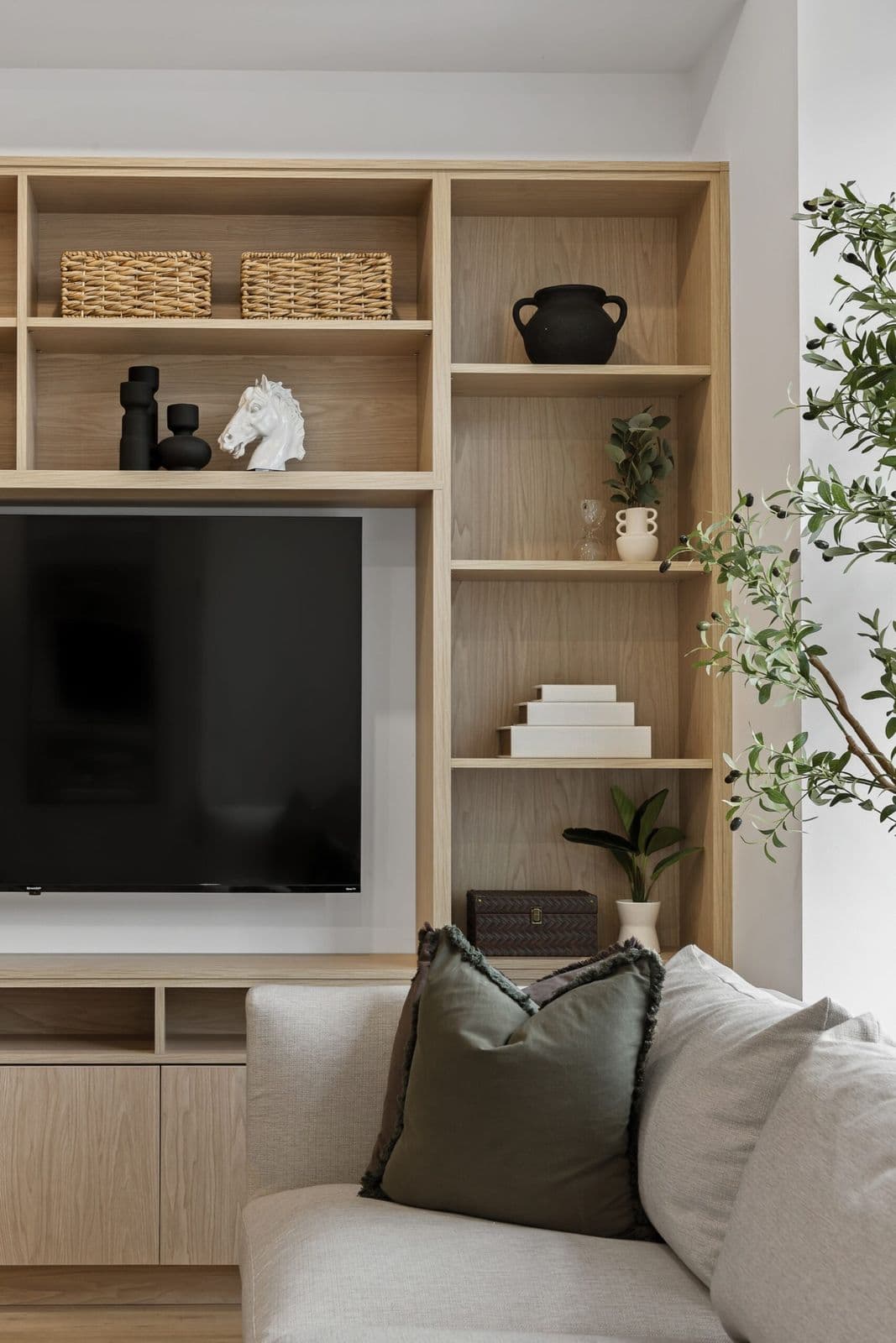Archer at Latimer – Living room built-in shelving with a wall-mounted TV, decorative vases and books, next to a sofa and an olive tree.
