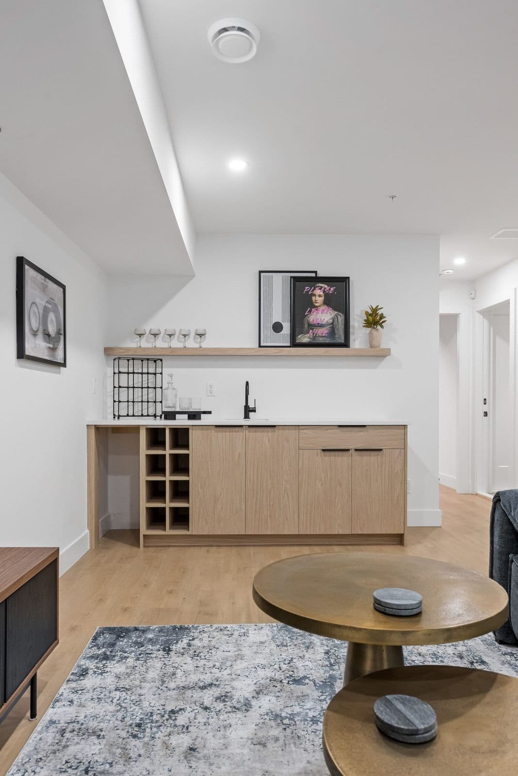 Archer at Latimer – Basement wet bar with wood cabinetry, wine rack, floating shelf with glassware and framed art, next to a seating area.