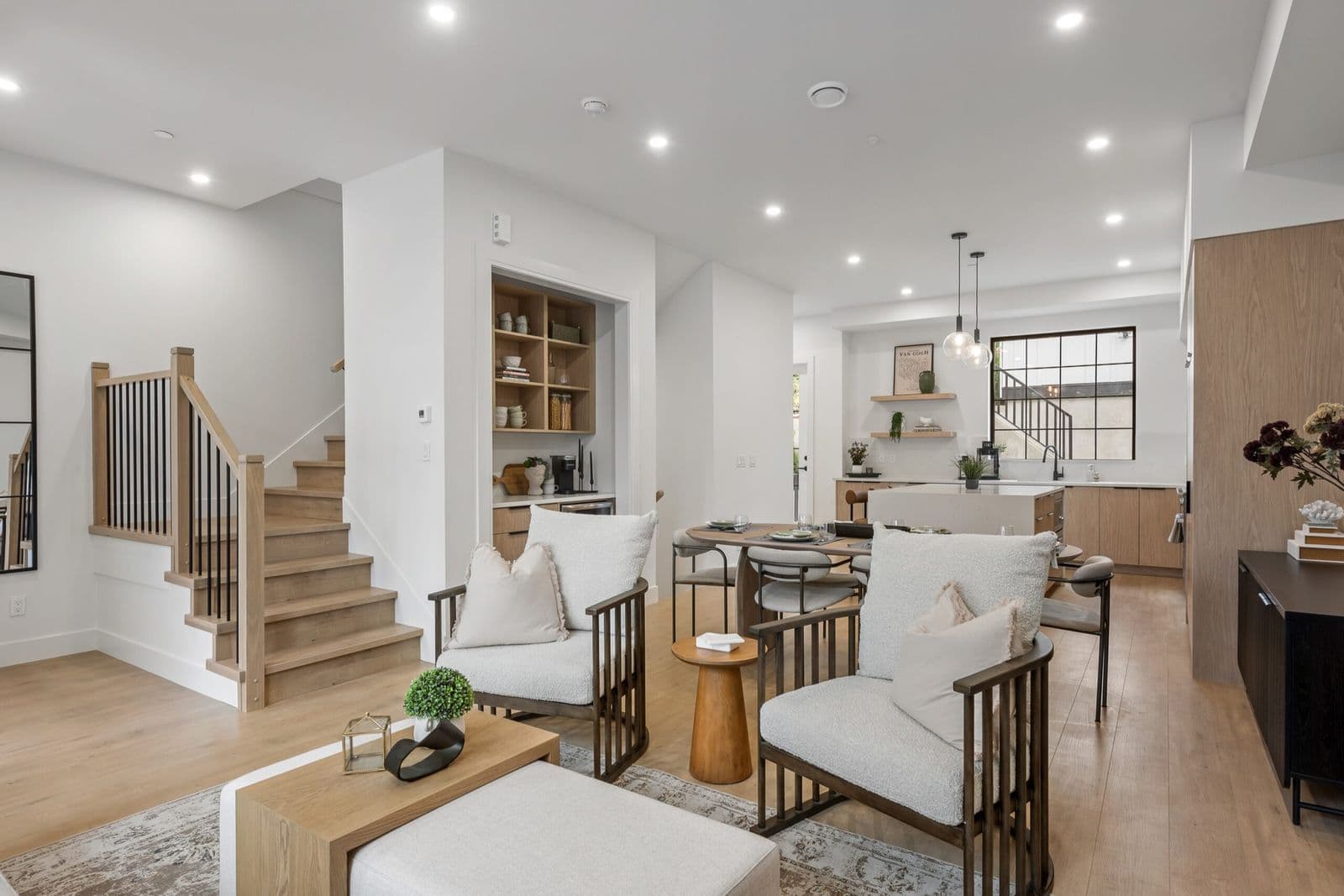 Archer at Latimer – Open-concept living room with wood-slat staircase, built-in shelving, and a dining/kitchen area visible beyond, light wood floors throughout.