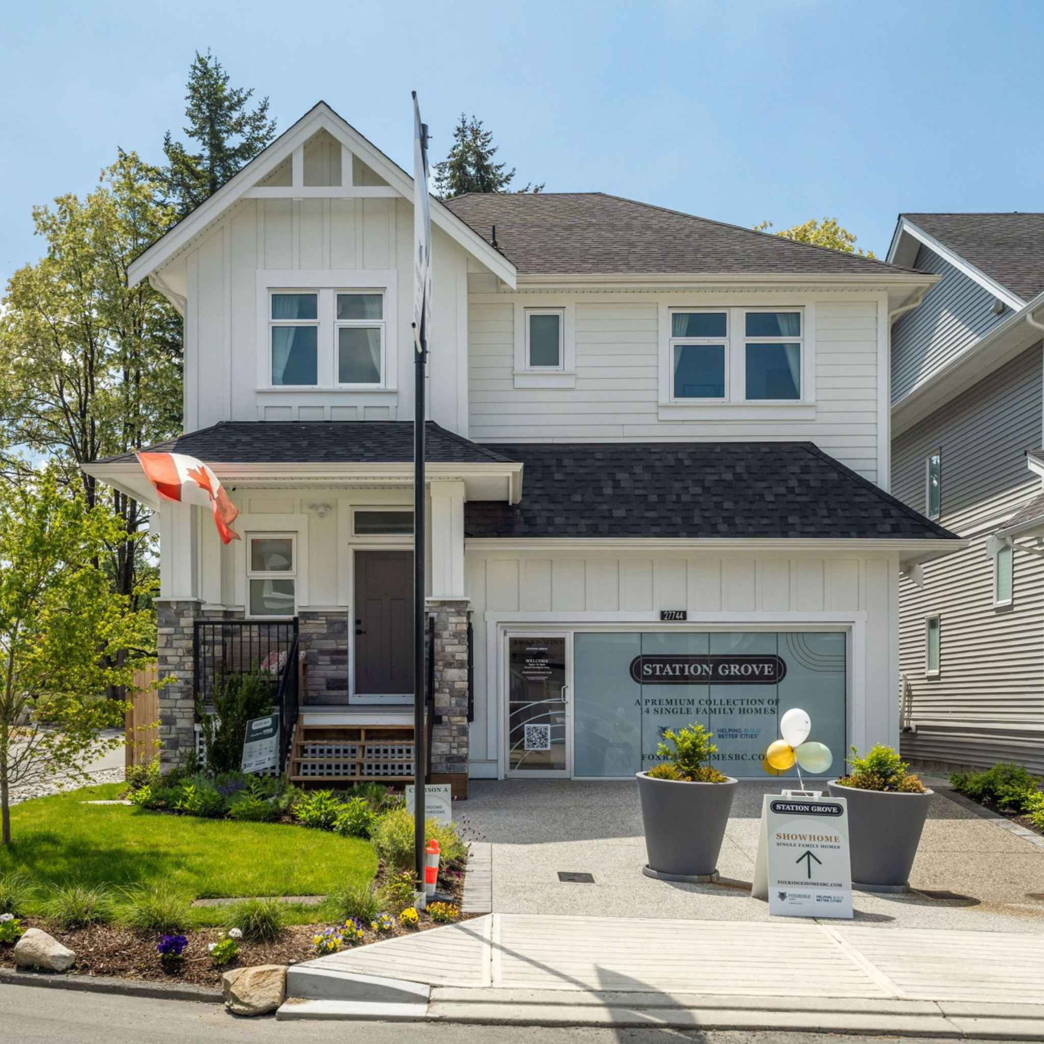 Station Grove – Front view of a white two-story showhome with a stone-clad porch, a Canadian flag, and a garage window branded 'Station Grove - A Premium Collection of Single Family Homes'.