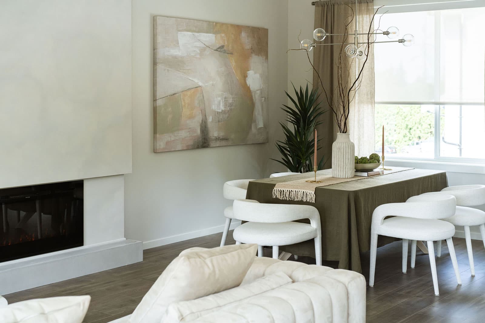 Fox and Aikins – Dining area with a fireplace feature wall, abstract painting, a table set with an olive tablecloth, and white upholstered chairs.