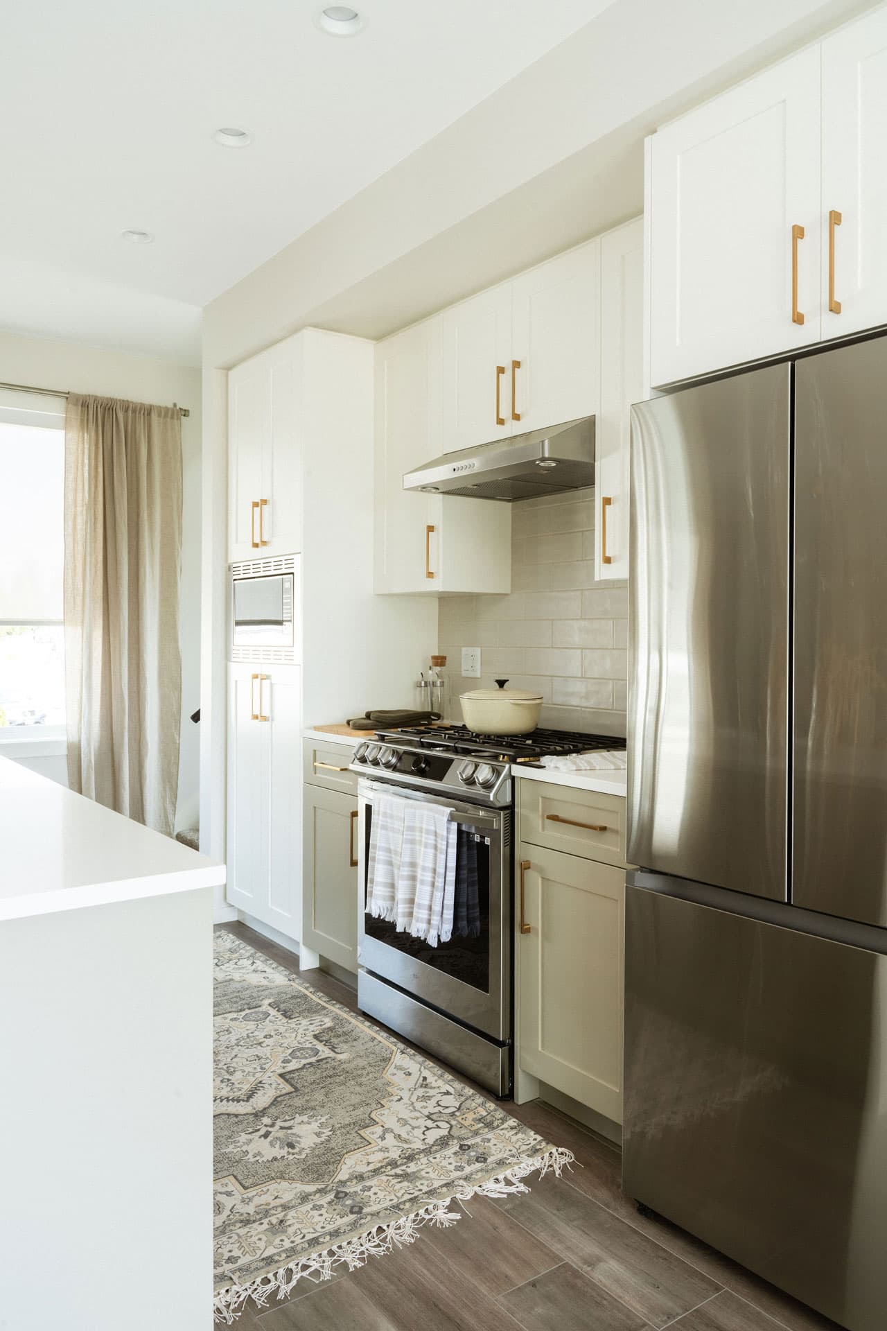 Fox and Aikins – Kitchen with white shaker cabinets, gold hardware, stainless steel range and refrigerator, and a patterned runner rug on the floor.