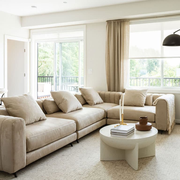 Fox and Aikins – Living room with a large beige sectional sofa, round coffee table, and sliding doors opening to a balcony with railing.