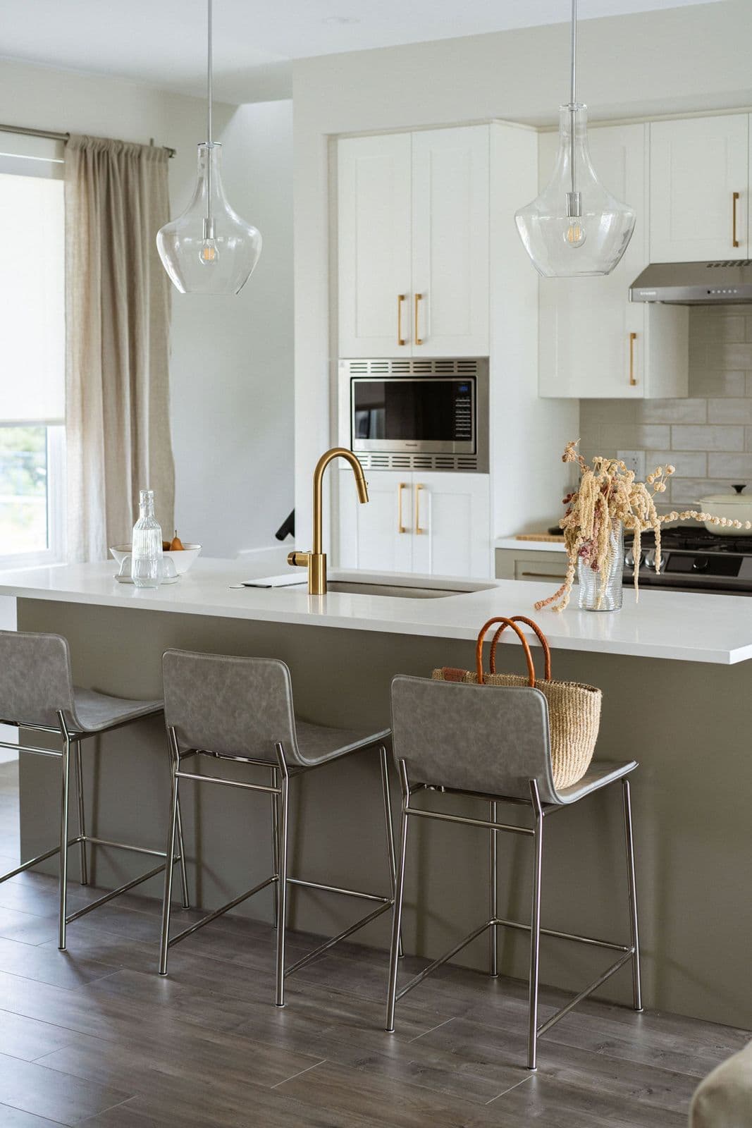 Fox and Aikins – Kitchen island with white quartz countertop, gold gooseneck faucet, glass pendant lights, and grey upholstered bar stools, white cabinetry behind.