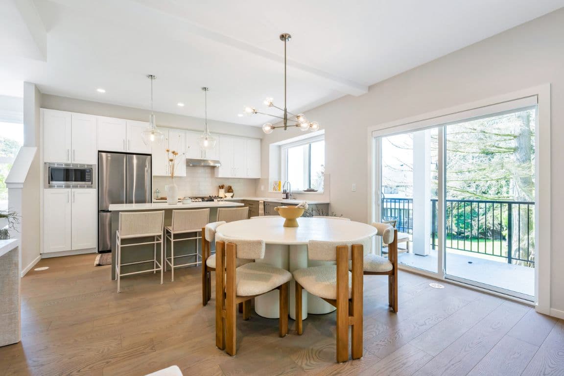 Fox and Aikins – Open-concept kitchen and dining area with a round table and wood chairs, pendant lighting, and sliding doors to a balcony with trees beyond.