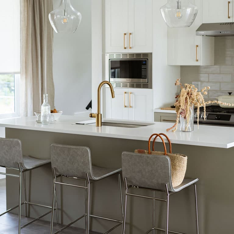 Fox and Aikins – Close-up of a kitchen island with white quartz countertop, gold faucet, glass pendant lights, and grey upholstered bar stools.