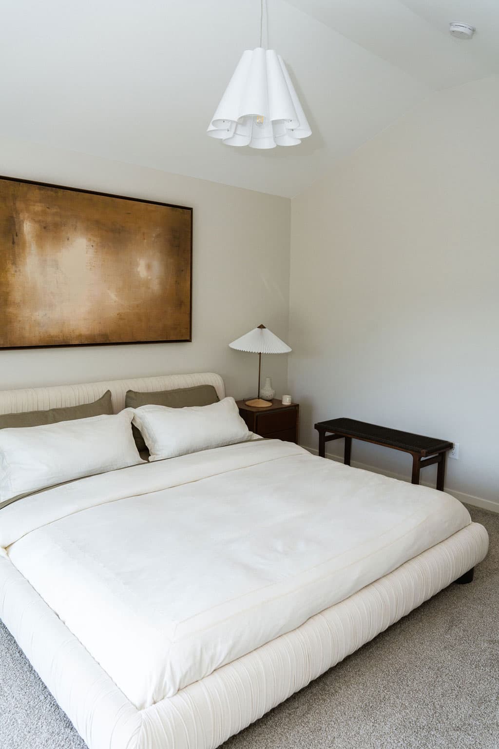 Fox and Aikins – Bedroom with a cream tufted bed, warm metallic abstract art above the headboard, and a pendant ceiling light.