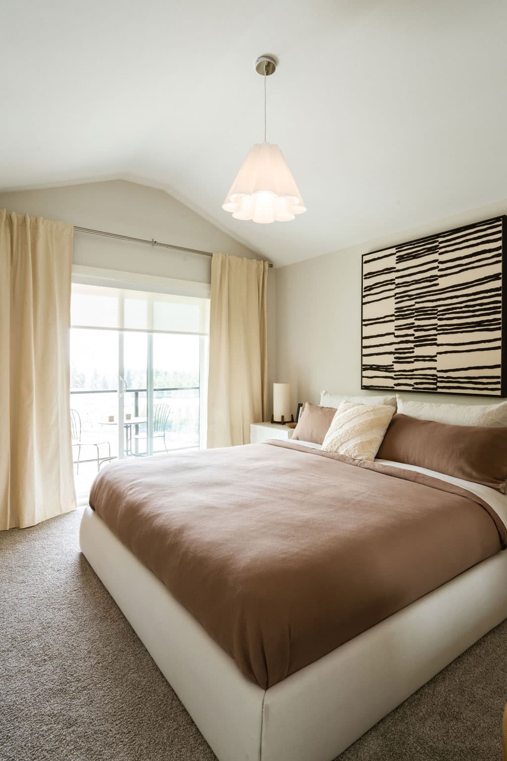 Fox and Aikins – Bedroom with a bed in taupe and cream bedding, pendant light fixture, and sliding glass doors opening to a balcony with a table and chairs.