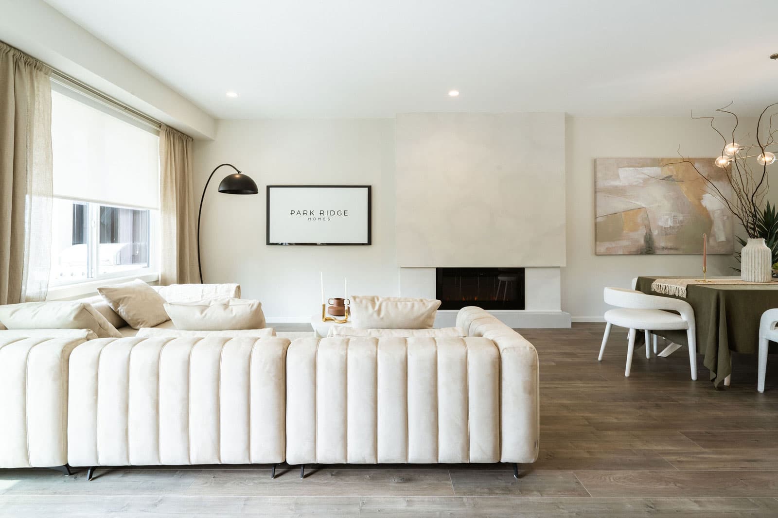 Fox and Aikins – Living room with a cream channel-tufted sofa facing a fireplace, a framed 'Park Ridge Homes' sign above a wall-mounted TV, dining area beyond.