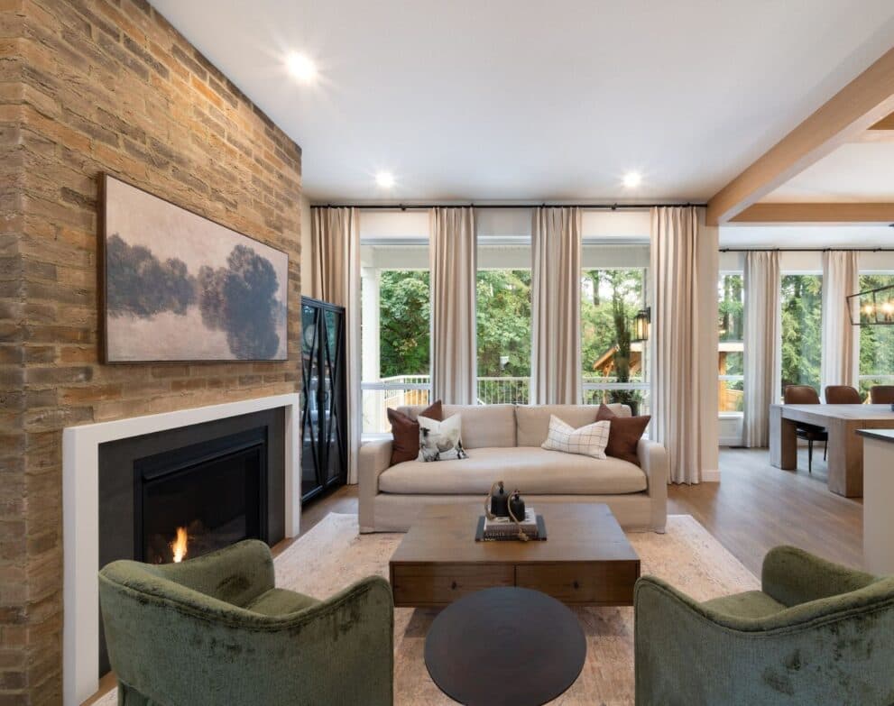 West Field at Cooper Meadows – Living room with a brick fireplace surround, framed landscape art, beige sofa with patterned throw pillows, and green accent armchairs, opening to a dining area and deck.