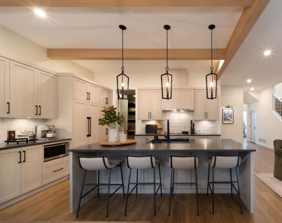 West Field at Cooper Meadows – Kitchen with cream shaker cabinetry, dark countertops, exposed wood ceiling beams, black cage pendant lights over an island with upholstered bar stools.