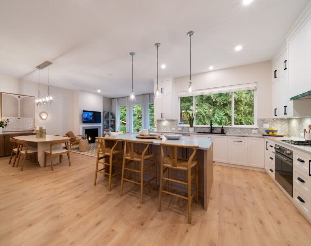 West Field at Cooper Meadows – Open-concept kitchen, dining and living area with white cabinetry, an island with wood barstools, a gas fireplace, and large windows overlooking trees.