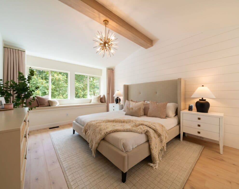 West Field at Cooper Meadows – Primary bedroom with a vaulted ceiling and exposed beam, sunburst chandelier, upholstered bed with neutral bedding, a window seat nook, and matching white nightstands.