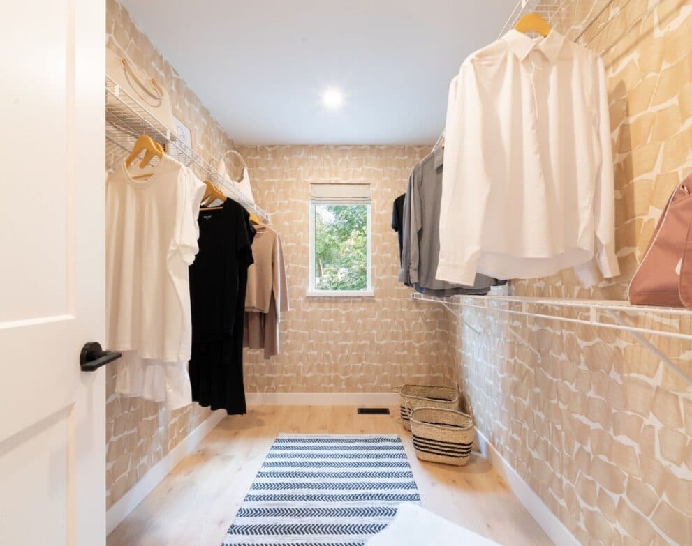 West Field at Cooper Meadows – Walk-in closet with patterned wallpaper, wire shelving holding folded clothes, hanging shirts and dresses, a window at the far end, and woven storage baskets.