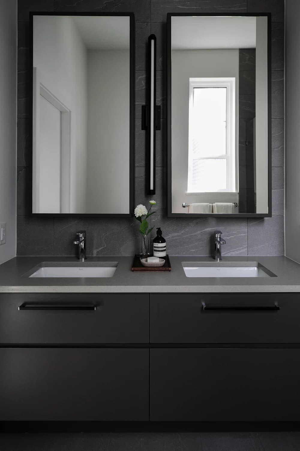 Cavendish – A modern bathroom double vanity featuring dark matte cabinetry, a light gray countertop, and dual undermount sinks with polished chrome faucets. Two tall, black-framed rectangular mirrors are mounted against a dark slate-gray tiled wall, separated by a sleek vertical light fixture.