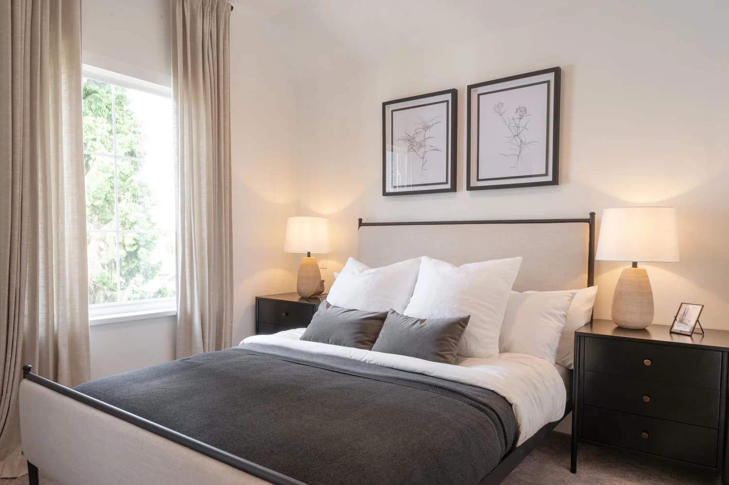 Cavendish – A warmly lit, cozy bedroom features a neatly made bed with white linens and a dark gray blanket flanked by matching black nightstands with glowing table lamps. Natural daylight filters through a large window with soft beige drapes, complementing the botanical framed artwork on the neutral wall.