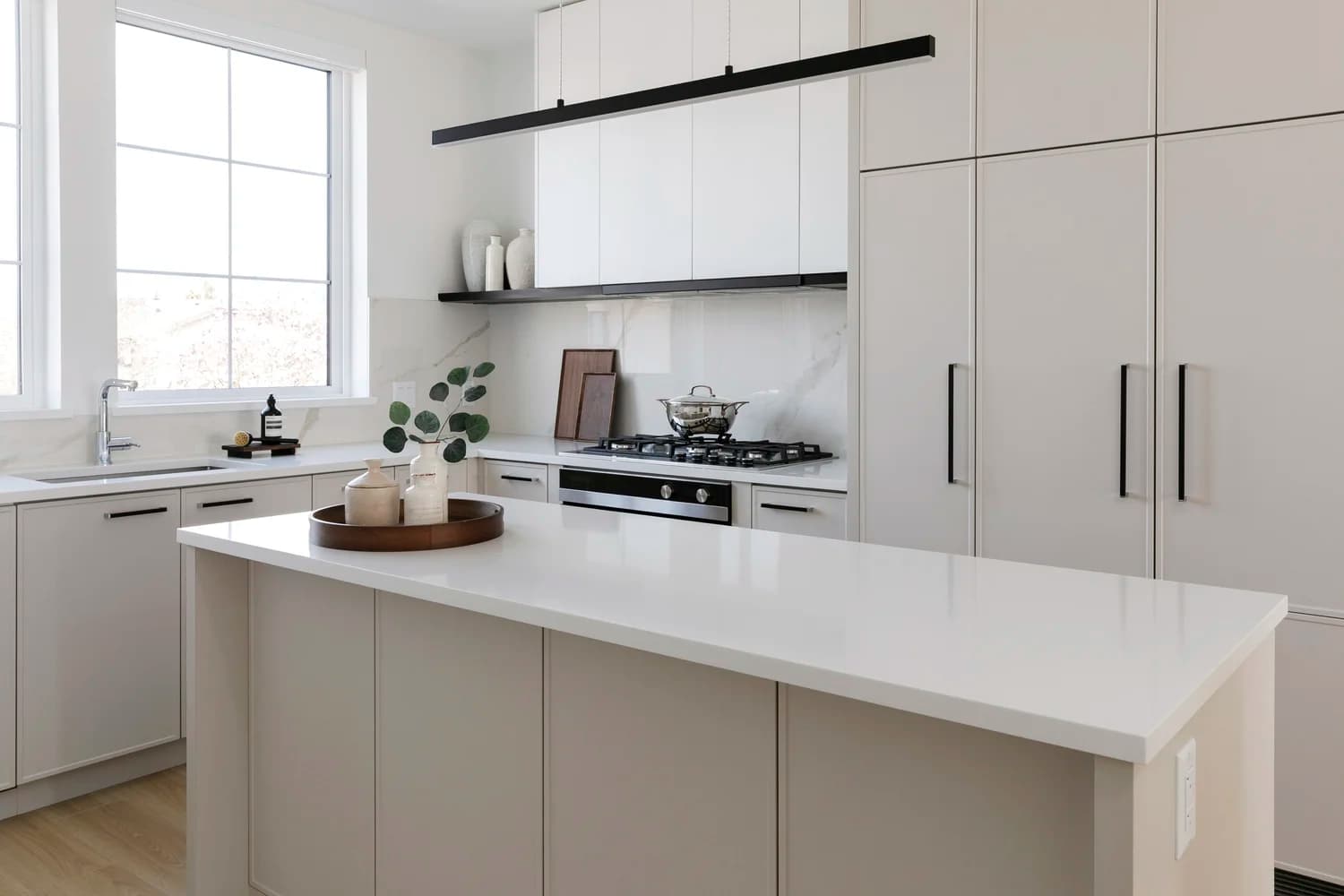 Cavendish – A bright, modern kitchen featuring off-white cabinetry with slim black handles, white quartz countertops, and a central island with decorative pottery. Natural daylight pours in through a large window next to the undermount sink, highlighting the marble backsplash and integrated stainless steel gas cooktop.