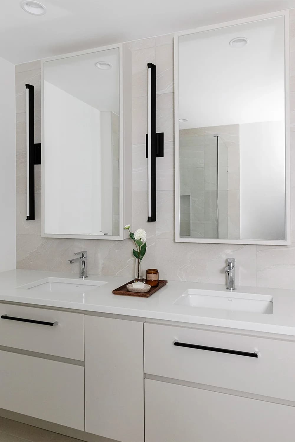 Cavendish – A bright, modern bathroom featuring a double vanity with white cabinetry, sleek black handles, and polished chrome faucets. Two large vertical rectangular mirrors are flanked by slim black light fixtures against a backdrop of soft beige stone-look wall tiles.