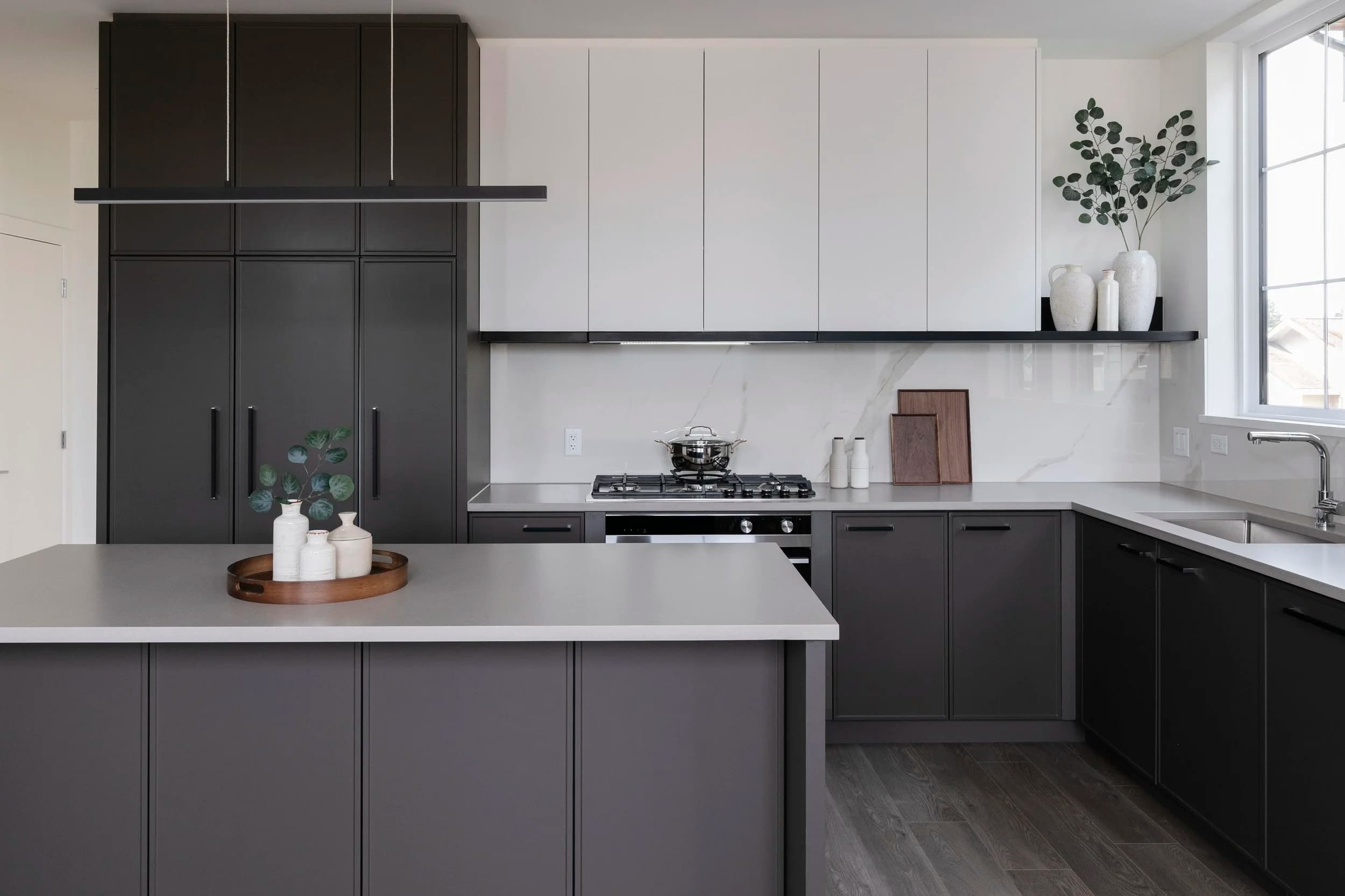 Cavendish – A modern kitchen featuring matte charcoal gray lower cabinetry and island paired with seamless white upper cabinets and a white marble-look backsplash. Light gray quartz countertops, a gas cooktop, and contemporary decor complete the space under bright natural light from an adjacent window.
