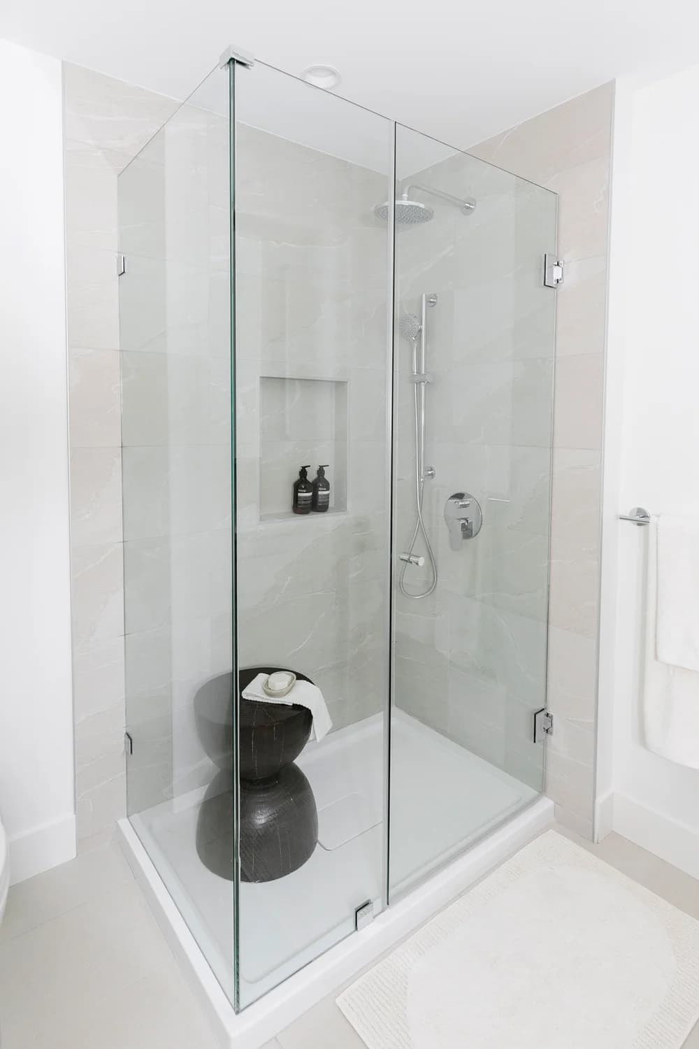 Cavendish – A modern walk-in shower enclosed in frameless glass features light gray marbled tiles, chrome fixtures including a rain showerhead and handheld wand, and a built-in recessed wall niche. Inside, a sculptural dark hourglass stool is styled with a white washcloth and soap bar, creating a clean, bright, and contemporary spa-like aesthetic.