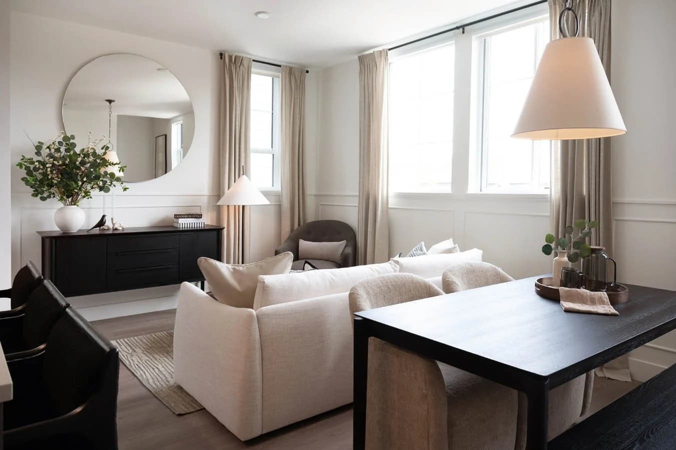 Cavendish – A bright, open-concept living and dining room filled with natural light from multiple large windows dressed in floor-length neutral curtains. The elegantly styled space features a cream fabric sofa, a dark wood dining table, textured accent chairs, and a large round mirror above a black sideboard.