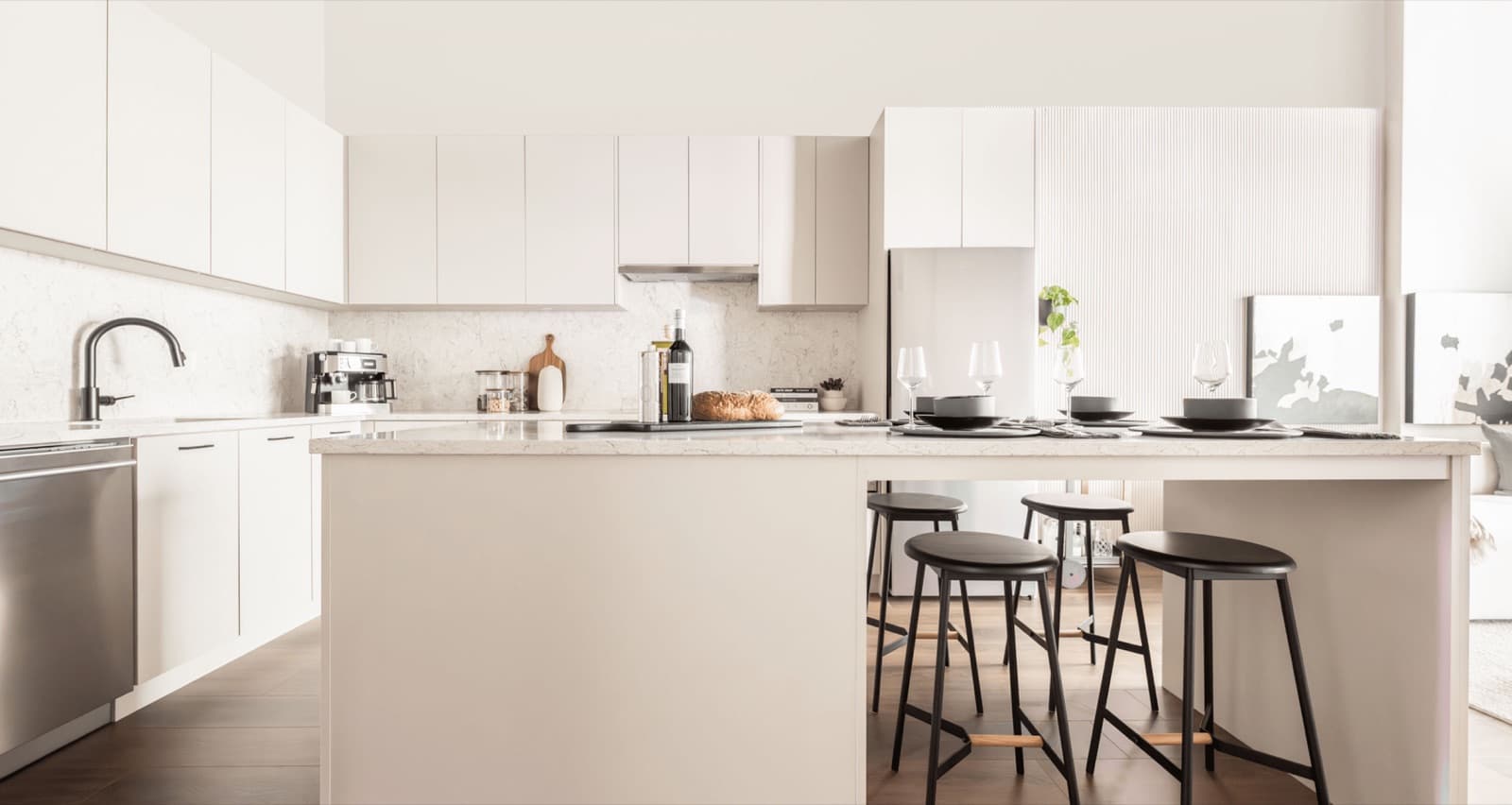 Rowan – A white kitchen with an island, black stools, wine and glassware set out, and a marble backsplash.
