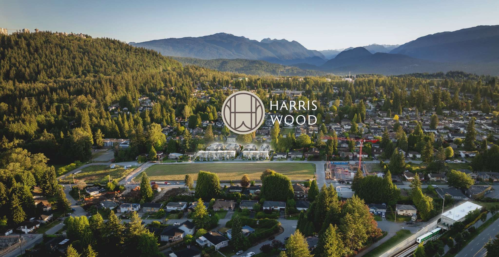 Harriswood – Drone view over a forested valley with mountains behind, showing the Harriswood townhome site under construction amid surrounding suburban houses, with the project logo overlaid.