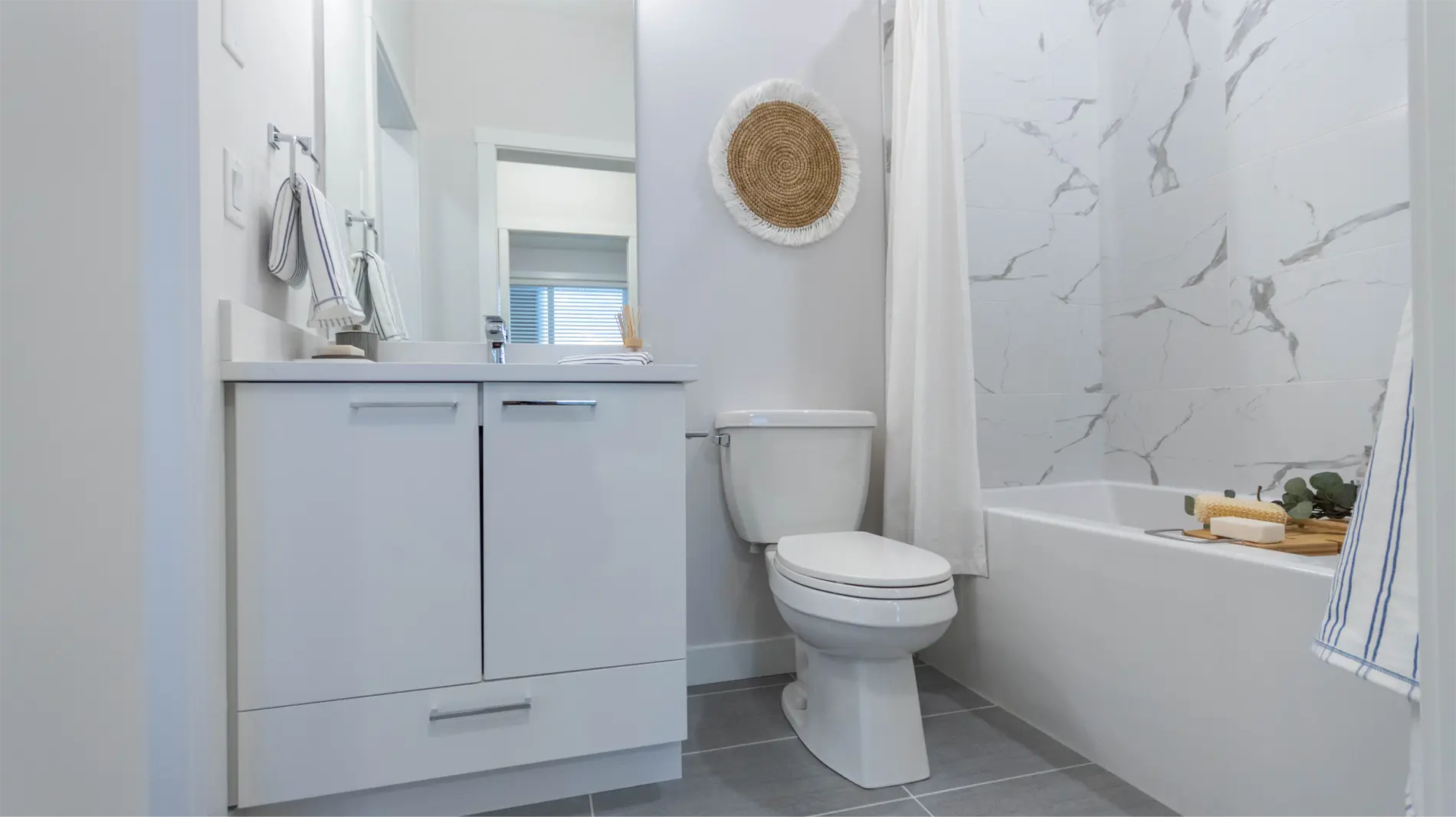 Montgomery – A white bathroom with a vanity, woven wall decor, a shower curtain over a tub, and grey tile flooring.