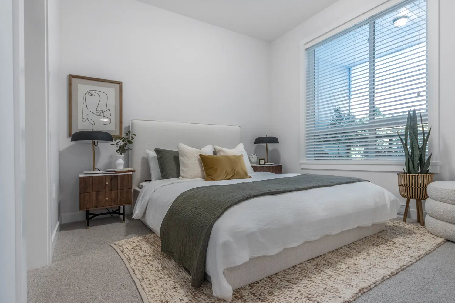 Montgomery – A bedroom with a white upholstered headboard, wood nightstands, green and mustard throw pillows, and a large window with blinds.