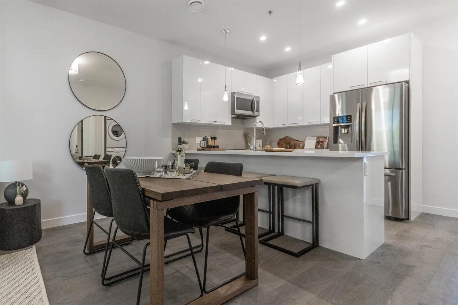 Montgomery – An open kitchen and dining area with white gloss cabinets, a stainless steel refrigerator, pendant lighting, and a wood dining table with dark chairs.