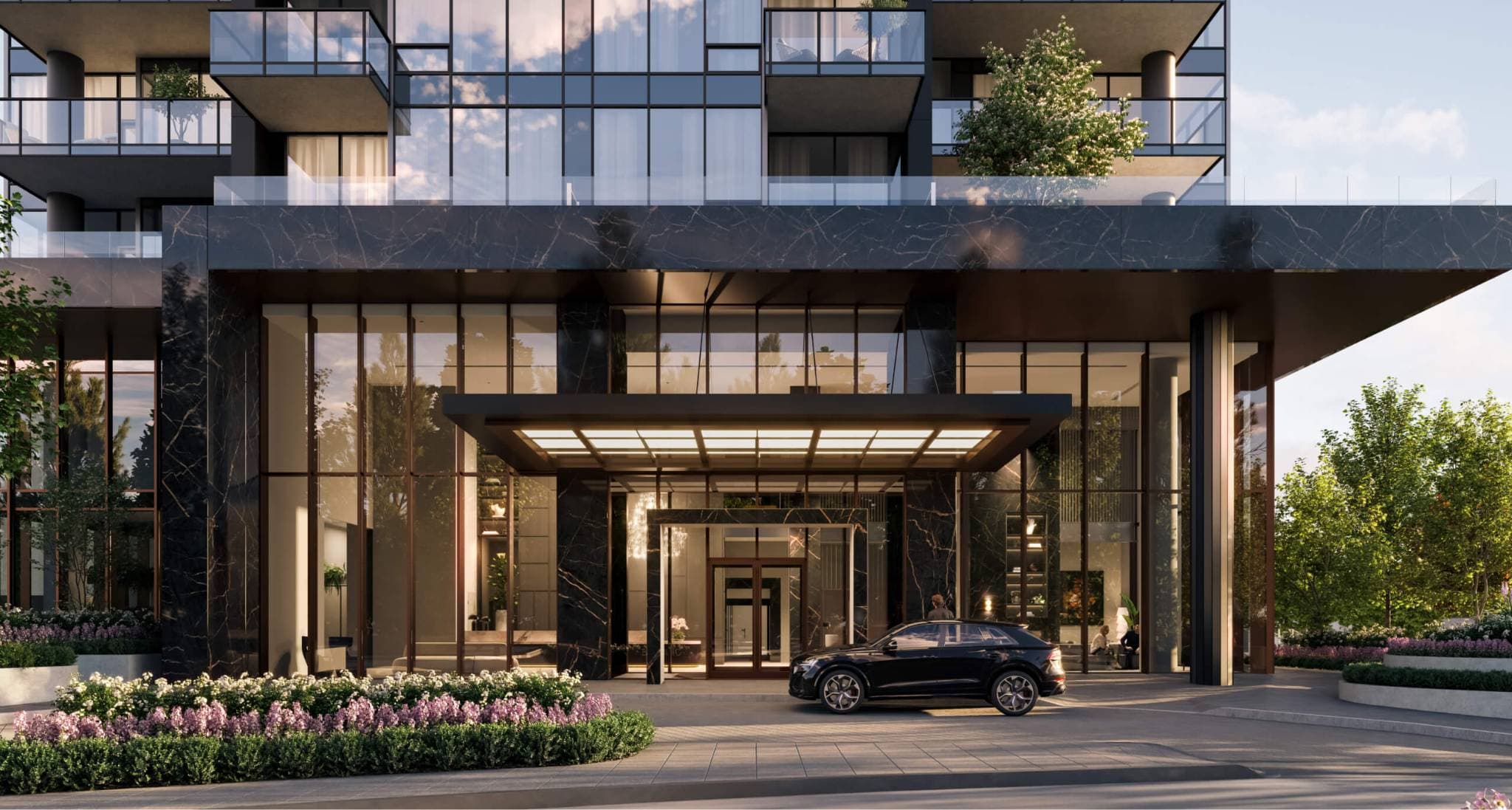 Bassano – A luxurious ground-level entrance to a modern high-rise featuring dark veined marble cladding, expansive glass walls, and an illuminated porte-cochère canopy. A black SUV is parked in the paved drop-off driveway, surrounded by manicured flower beds and lush green landscaping in daylight.