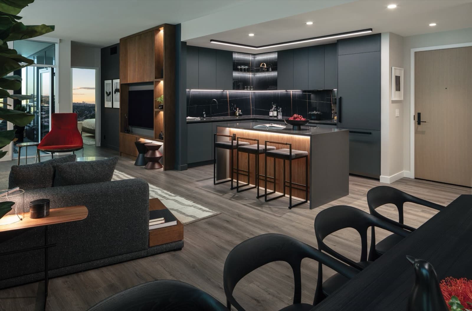 Bassano – Open-concept living and kitchen area with dark grey cabinetry, black marble backsplash, wood-accented kitchen island with bar stools, and a red accent chair by the window with a city view.