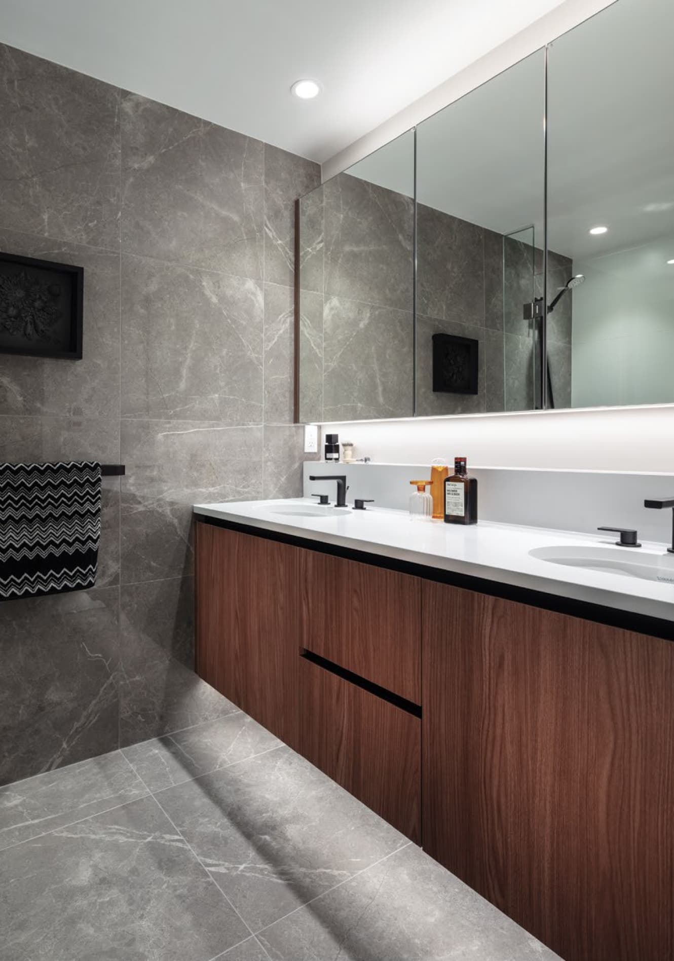 Bassano – Bathroom with a floating double-sink vanity in dark wood, grey marble-tile walls and floor, large mirror, and a walk-in shower visible in the mirror reflection.