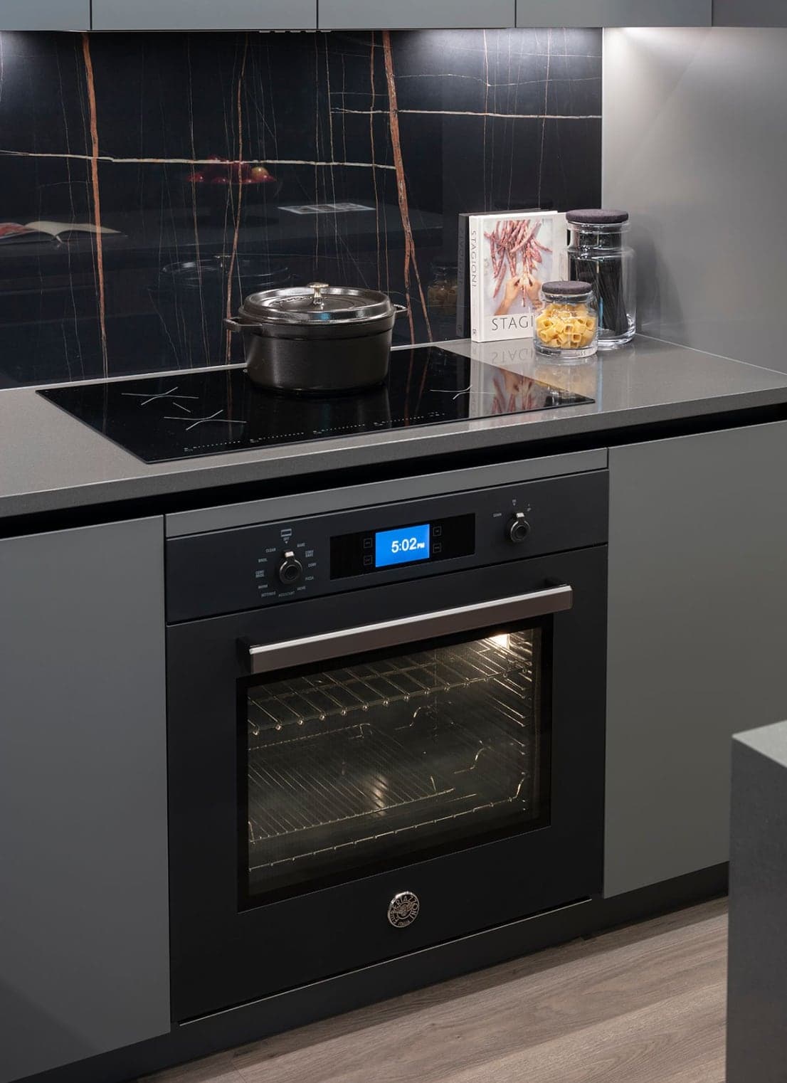 Bassano – Close-up of a built-in oven with digital display and induction cooktop holding a black dutch oven, dark marble backsplash behind.