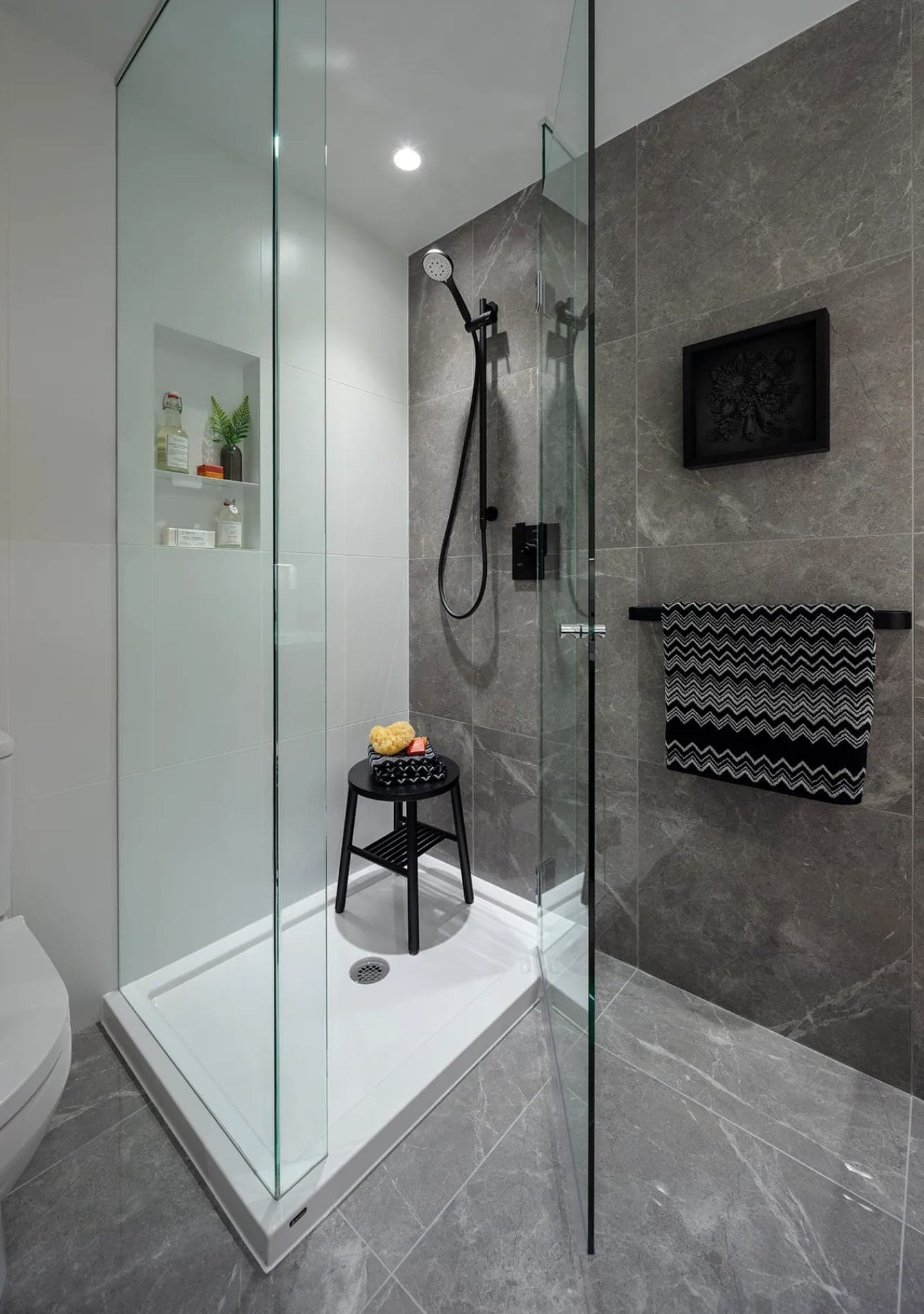 Bassano – Glass-enclosed walk-in shower with grey marble tile, black fixtures, a small black stool, and a patterned towel on a wall-mounted bar.
