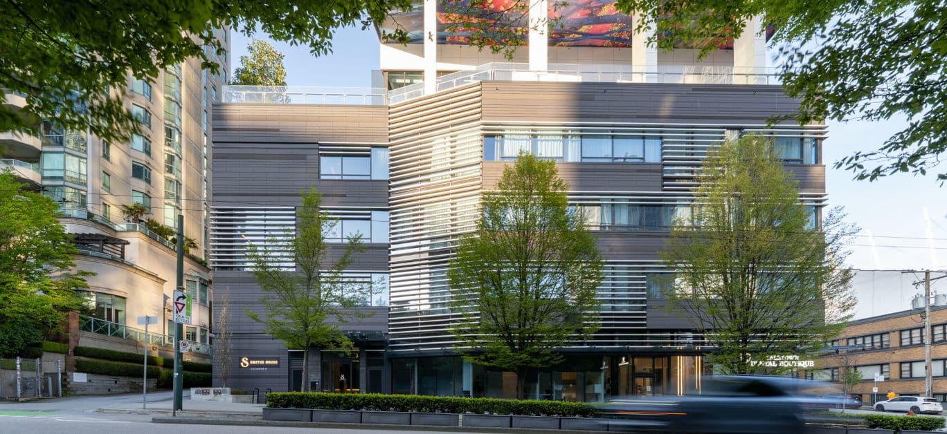 Bassano – Street-level photo of a modern multi-story building with dark metal cladding and louvered sunshades, trees in front, and visible signage reading 'Smithe House' and 'Downtown Dental Boutique' at ground level.