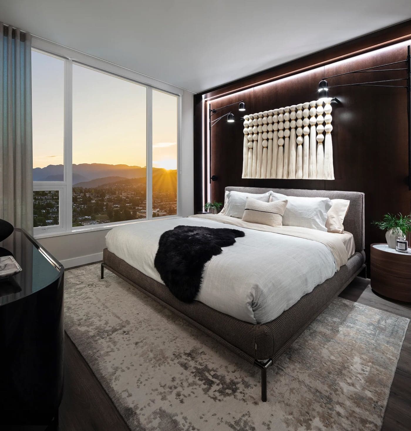 Bassano – Bedroom with a dark wood upholstered headboard wall, macrame wall hanging, a bed with a black fur throw, and a large window showing a sunset over hills.