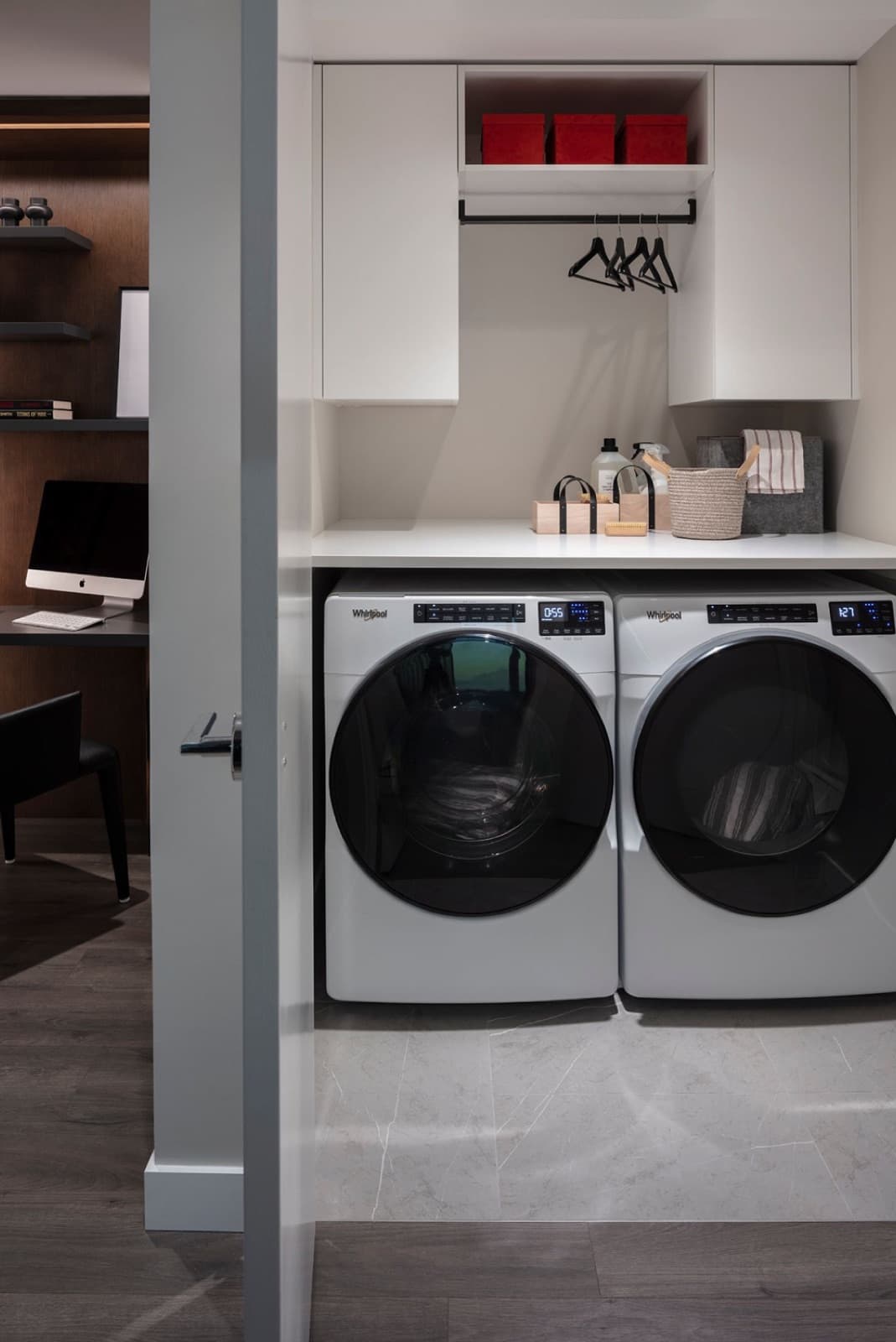 Bassano – In-suite laundry nook with a stacked white front-load washer and dryer, upper cabinet storage with hanging rod, next to a home office desk visible through the doorway.
