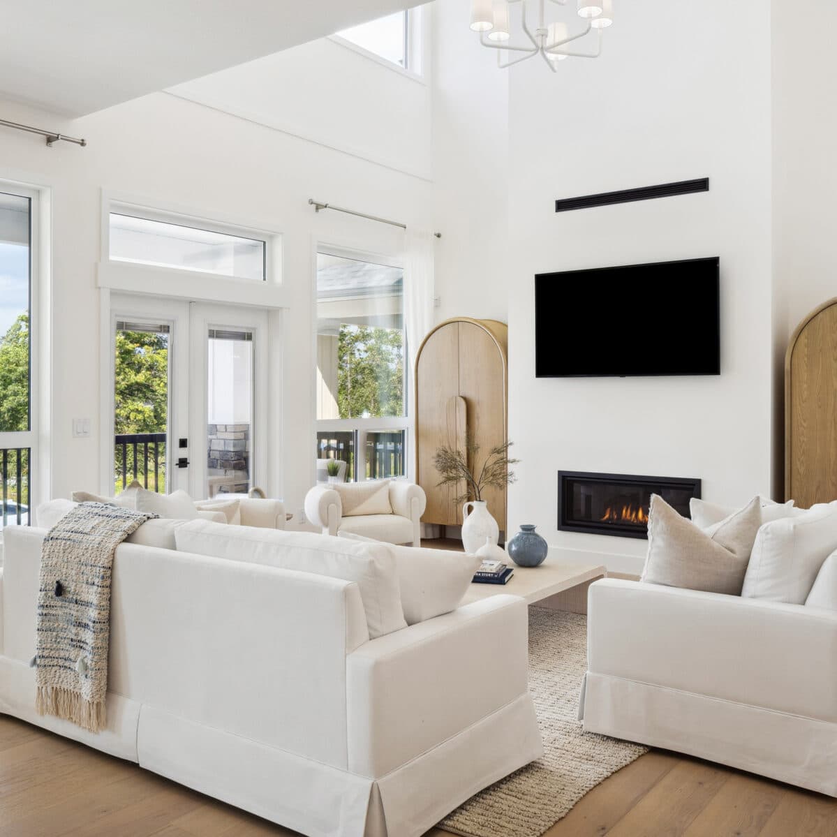 Abbey Heights – Bright double-height living room with white slipcovered sofas, a gas fireplace, wall-mounted TV, and large windows to a balcony.