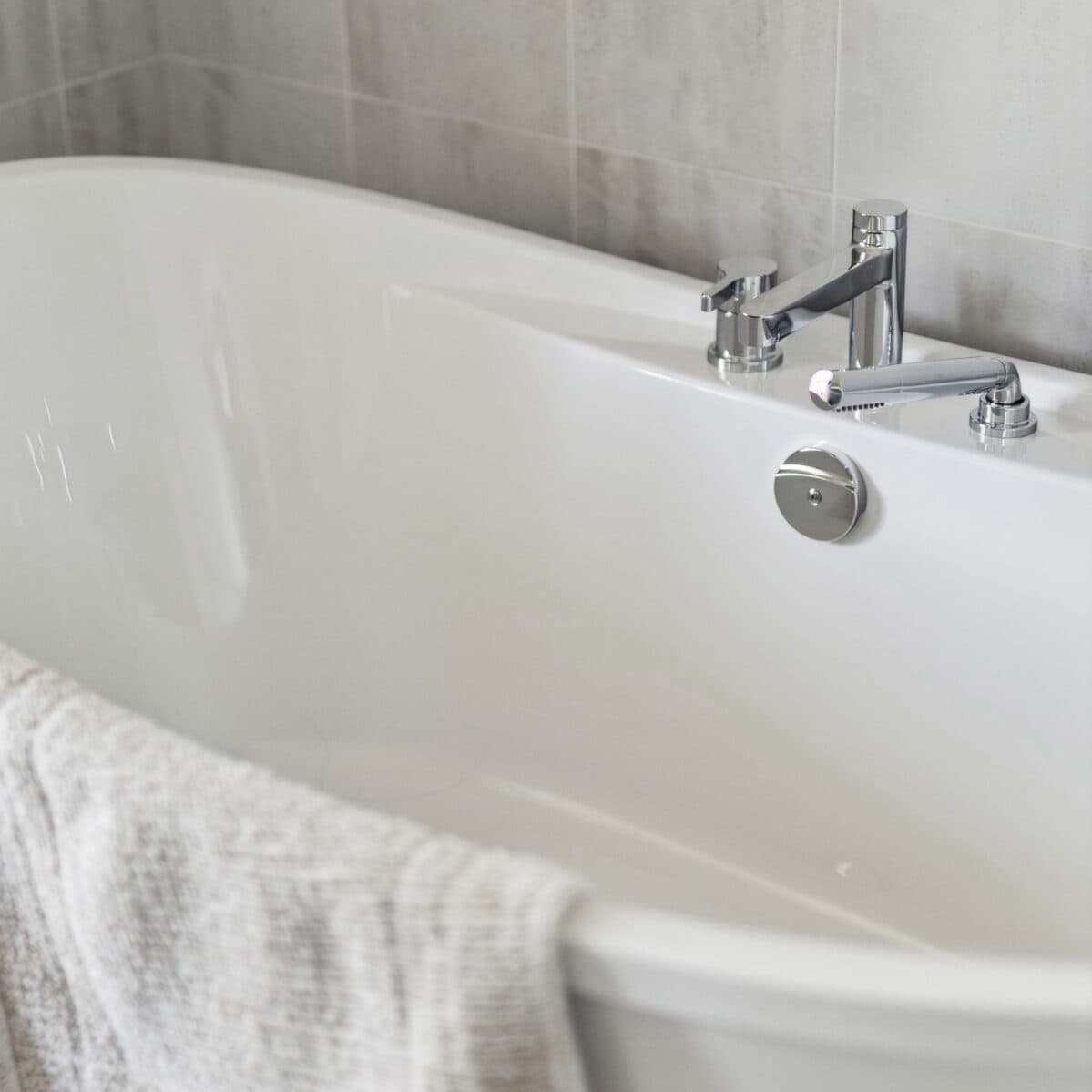 Abbey Heights – Close-up of a white soaker tub with chrome deck-mounted faucet and handheld sprayer against tiled walls.
