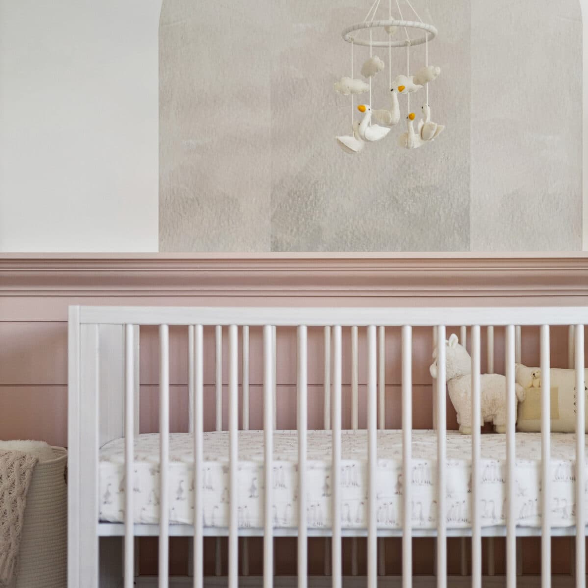 Abbey Heights – Close-up of a white crib in a nursery with pink wainscoting, a hanging bird mobile, and plush toys.