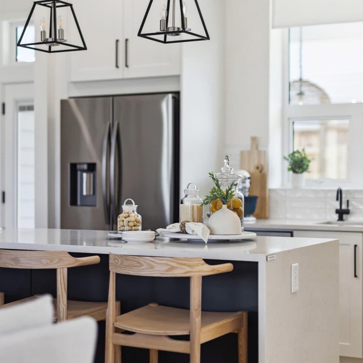 Abbey Heights – Kitchen island close-up with wood bar stools, black pendant lights, a stainless steel fridge, and decorative glass jars on the counter.