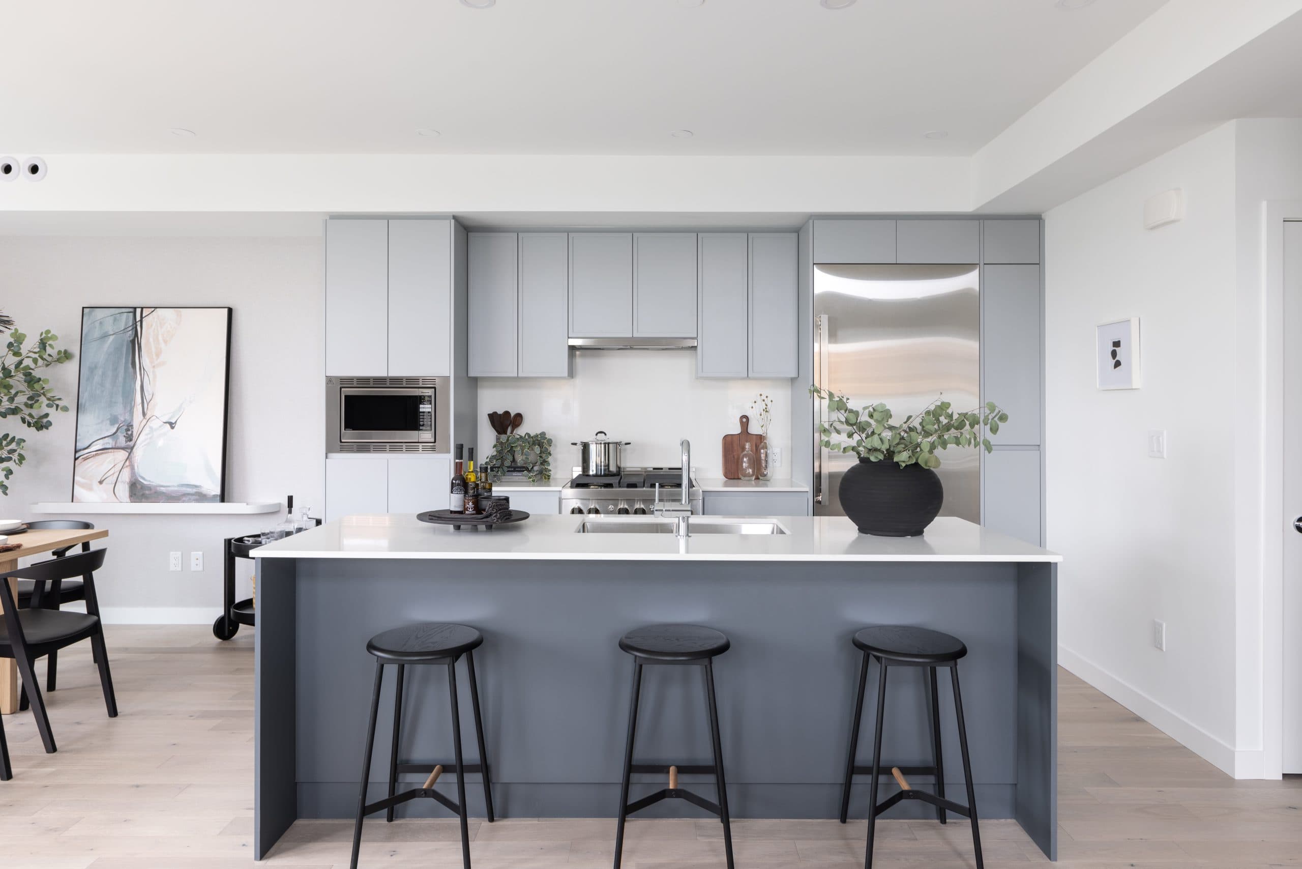 Ocean Row – A modern kitchen with light gray cabinetry, a large stainless refrigerator, a navy-blue waterfall-edge island with black stools, and light hardwood floors.