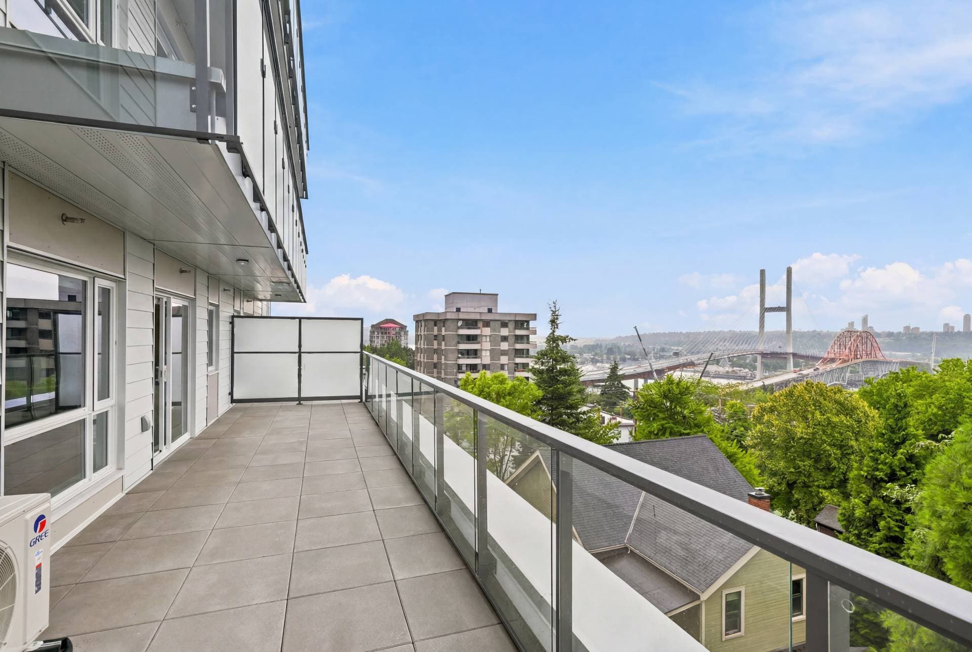 First & Royal – A spacious modern balcony with tiled flooring and glass railings offers sweeping daytime views of green treetops, surrounding residential buildings, and a large suspension bridge spanning a river in the distance. The building facade features clean white siding and glass sliding doors under a bright blue sky with light clouds.