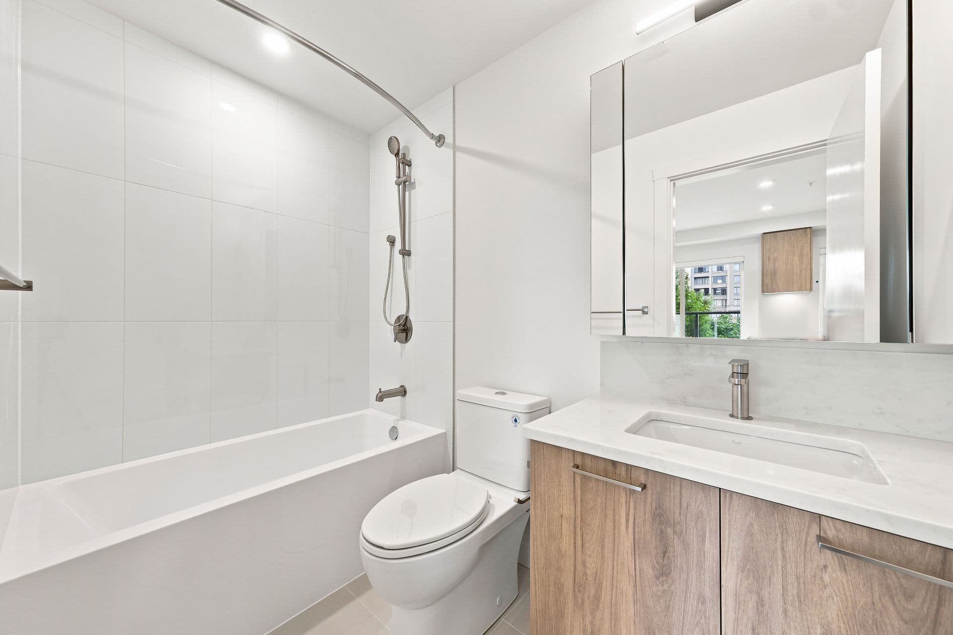 First & Royal – A modern, bright bathroom featuring a white soaking tub with a curved shower curtain rod, a white toilet, and a warm wood-grain vanity with a quartz countertop. Large white wall tiles and a mirrored medicine cabinet enhance the clean, contemporary aesthetic.