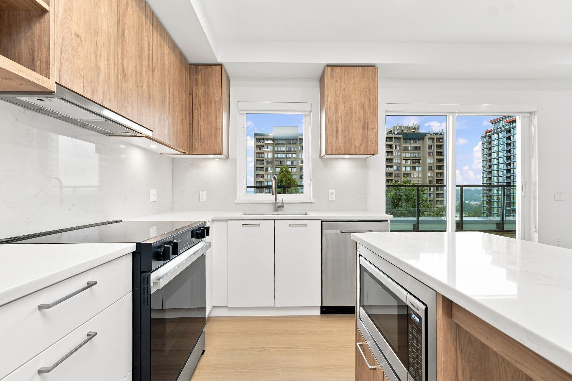 First & Royal – A modern kitchen featuring white quartz countertops, white lower cabinetry, light wood-grain upper cabinets, and integrated stainless steel appliances. A window above the sink and an adjacent glass sliding door look out onto neighboring residential high-rises under a clear blue sky.