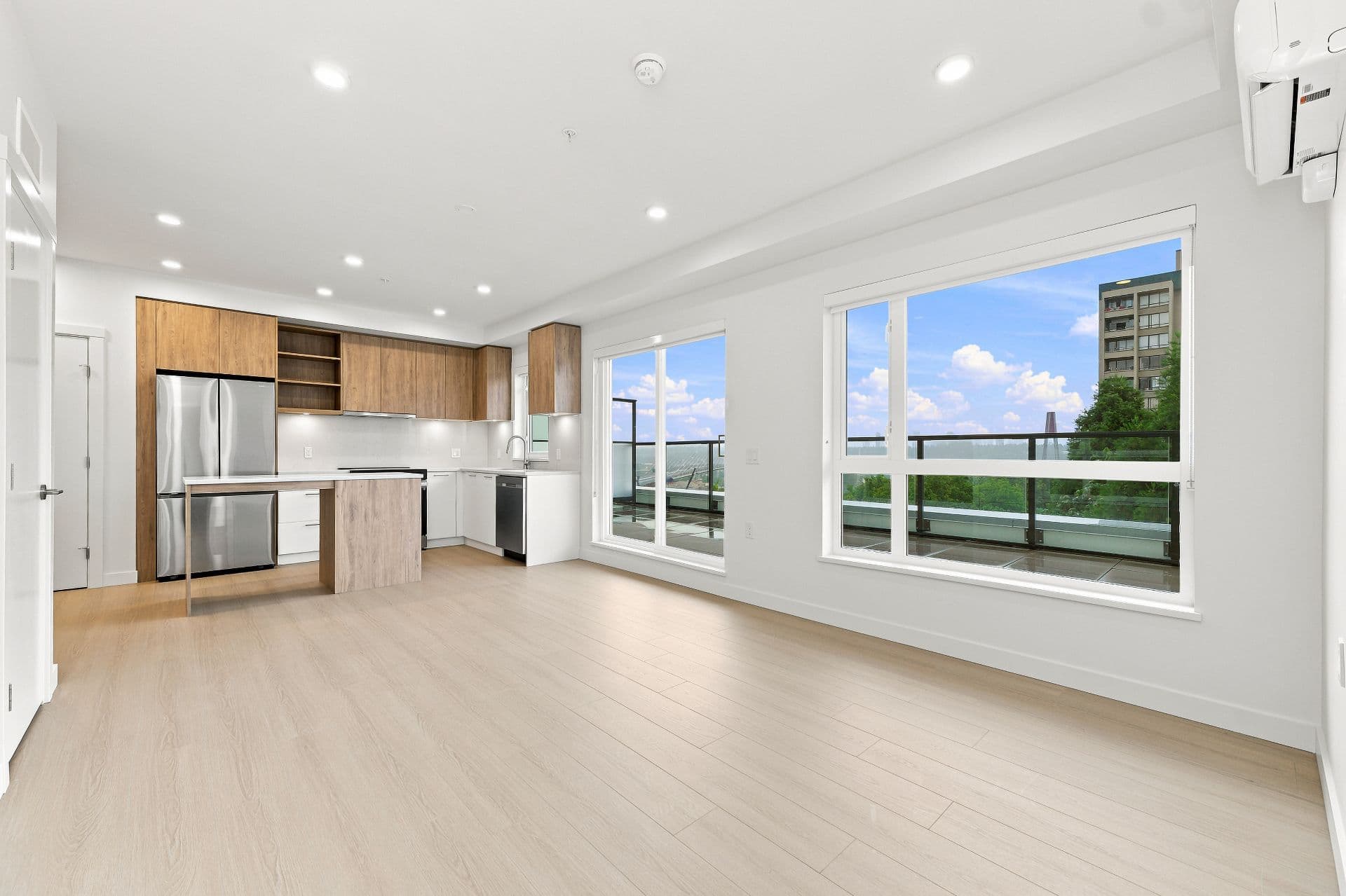 First & Royal – A bright, open-concept condo interior featuring light hardwood floors, white walls, and an L-shaped kitchen with warm wood upper cabinets, stainless steel appliances, and a small prep island. Large windows and a glass balcony door fill the empty living space with natural daylight, offering a view of a blue sky and distant trees.