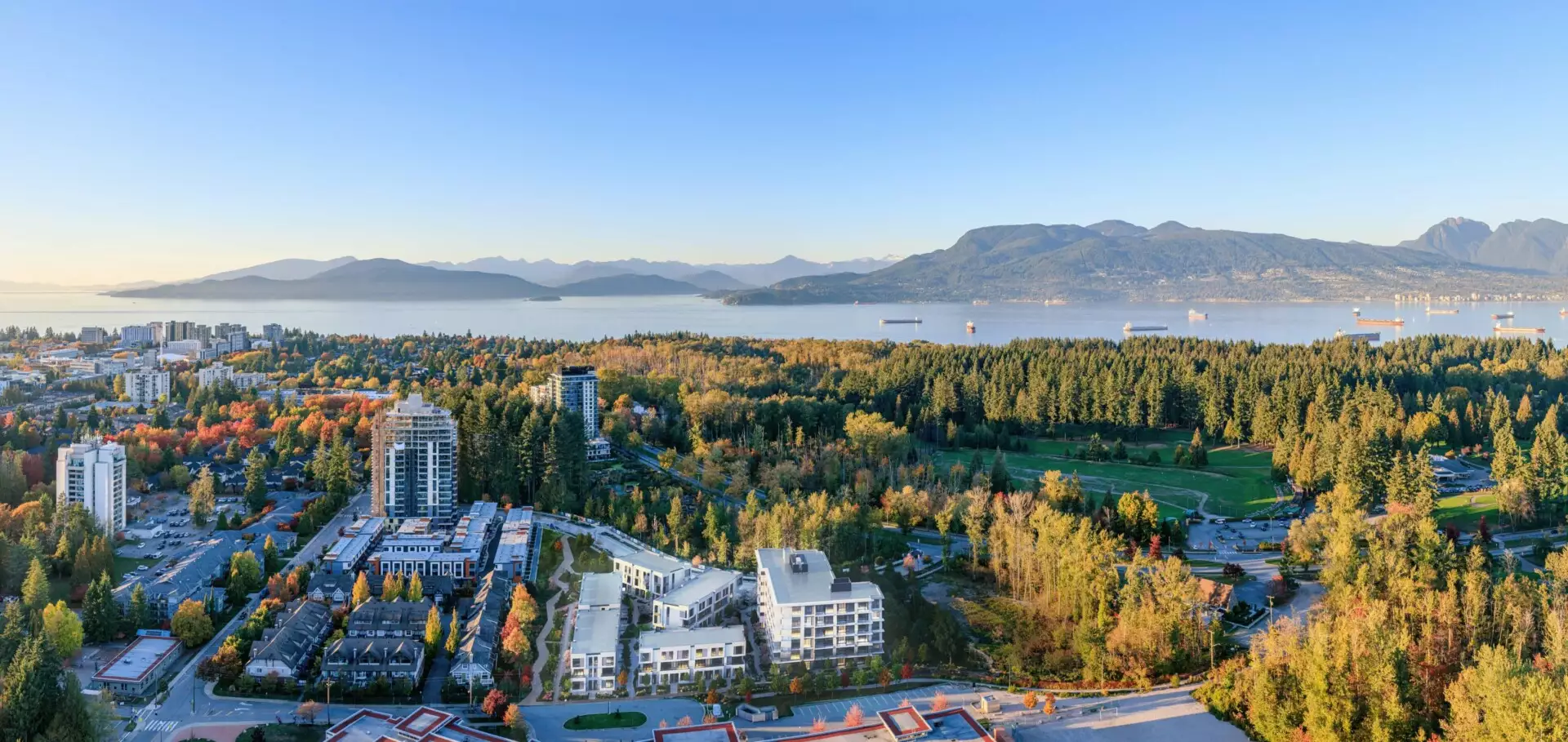 Edge Way at leləm̓ – A high-altitude aerial view overlooking residential developments and townhomes nestled among lush autumn-colored forests and a golf course. In the background, expansive blue waters dotted with cargo ships stretch toward distant mountain ranges under a clear, bright sky.
