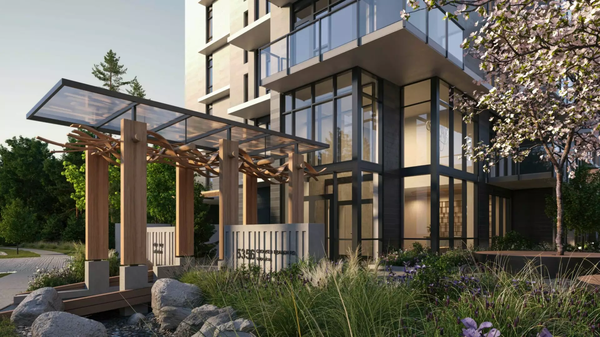 Edge Way at leləm̓ – A ground-level view of a contemporary mid-rise residential building entrance featuring double-height glass lobby windows and upper glass balconies. In the foreground, a modern timber pergola with a glass canopy spans a landscaped pathway surrounded by lush ornamental grasses, purple flowers, and a blooming tree in bright daylight.